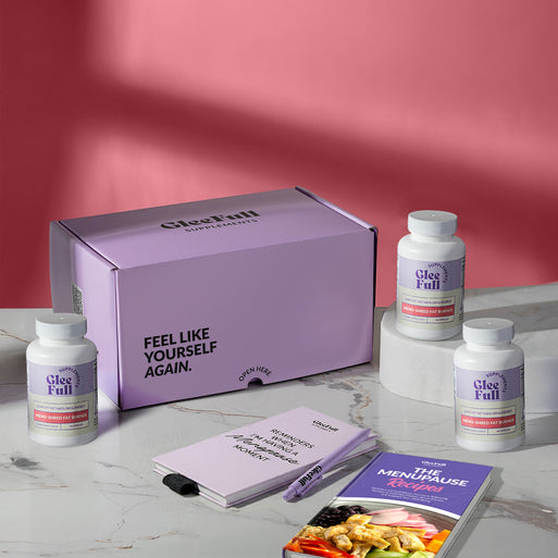 Subscription bundle display for GleeFull Meno-Shred on a marble surface against a pink background. The layout includes a large purple shipping box reading 'FEEL LIKE YOURSELF AGAIN.' and 'OPEN HERE', three white bottles of Meno-Shred Fat Burner, a purple branded pen, a matching notepad that reads 'REMINDERS WHEN I'M HAVING A MENOPAUSE MOMENT', and a recipe booklet titled 'THE MENOPAUSE Recipes'.