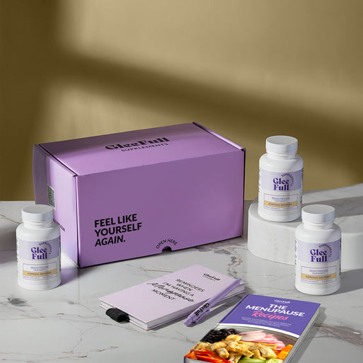 A product bundle display for GleeFull Supplements on a marble surface against a yellow background. The layout includes a large purple shipping box featuring the slogan 'FEEL LIKE YOURSELF AGAIN.', three white bottles of Adrenal Restore, a purple branded pen, a matching notepad that reads 'REMINDERS WHEN I'M HAVING A MENOPAUSE MOMENT', and a recipe booklet titled 'THE MENOPAUSE Recipes'