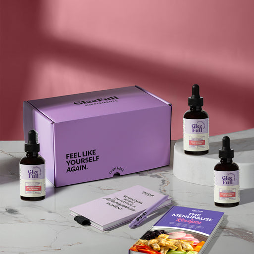 Subscription bundle display for GleeFull Meno Morning on a marble surface against a pink background. The layout includes a large purple shipping box reading 'FEEL LIKE YOURSELF AGAIN.' and 'OPEN HERE', three amber dropper bottles of Meno Morning Fat Burner, a purple branded pen, a matching notepad that reads 'REMINDERS WHEN I'M HAVING A MENOPAUSE MOMENT', and a recipe booklet titled 'THE MENOPAUSE Recipes'.