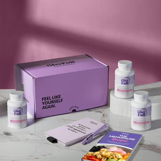 A product bundle display for GleeFull Supplements on a marble surface against a pink background. The layout includes a large purple shipping box featuring the slogan 'FEEL LIKE YOURSELF AGAIN.', three white bottles of Thyroid Balance, a purple branded pen, a matching notepad that reads 'REMINDERS WHEN I'M HAVING A MENOPAUSE MOMENT', and a recipe booklet titled 'THE MENOPAUSE Recipes'