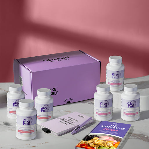 A product bundle display for GleeFull Supplements on a marble surface against a red background. The layout includes a large purple shipping box featuring the slogan 'FEEL LIKE YOURSELF AGAIN.', three white bottles of Meno Shred Fat Burner and three white bottler of Over 30 Hormone Support, a purple branded pen, a matching notepad that reads 'REMINDERS WHEN I'M HAVING A MENOPAUSE MOMENT', and a recipe booklet titled 'THE MENOPAUSE Recipes'