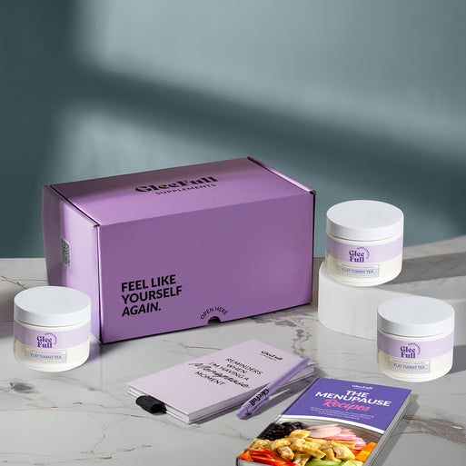 A product bundle display for GleeFull Supplements on a marble surface against a blue background. The layout includes a large purple shipping box featuring the slogan 'FEEL LIKE YOURSELF AGAIN.', three white bottles of Flat Tummy Tea, a purple branded pen, a matching notepad that reads 'REMINDERS WHEN I'M HAVING A MENOPAUSE MOMENT', and a recipe booklet titled 'THE MENOPAUSE Recipes'
