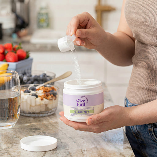 A person scoops white powder from a GleeFull Meno Creatine jar over a glass on a marble countertop, with a yogurt bowl, water glass and fresh strawberries beside it. Label reads 'Muscle & Joint Support, Meno Creatine'.