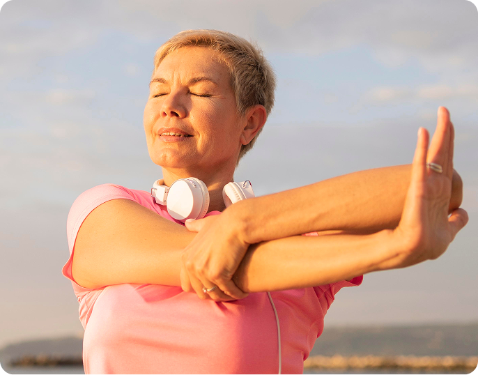 A woman outdoors stretching her arm, wearing a pink athletic top and white over-ear headphones around her neck, with a soft-focus sky and water background.